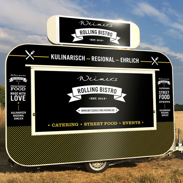 Schwarz-gold beklebter Verkaufsanhänger „Weimers Rolling Bistro“ auf einem Feld unter blauem Himmel.